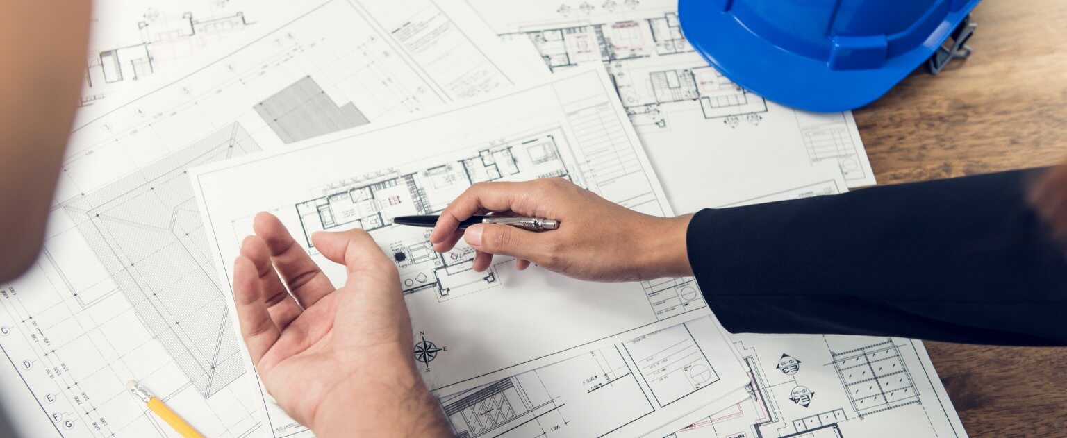 Hands point with a pen at architectural blueprints spread on a wooden desk, alongside a blue hard hat.