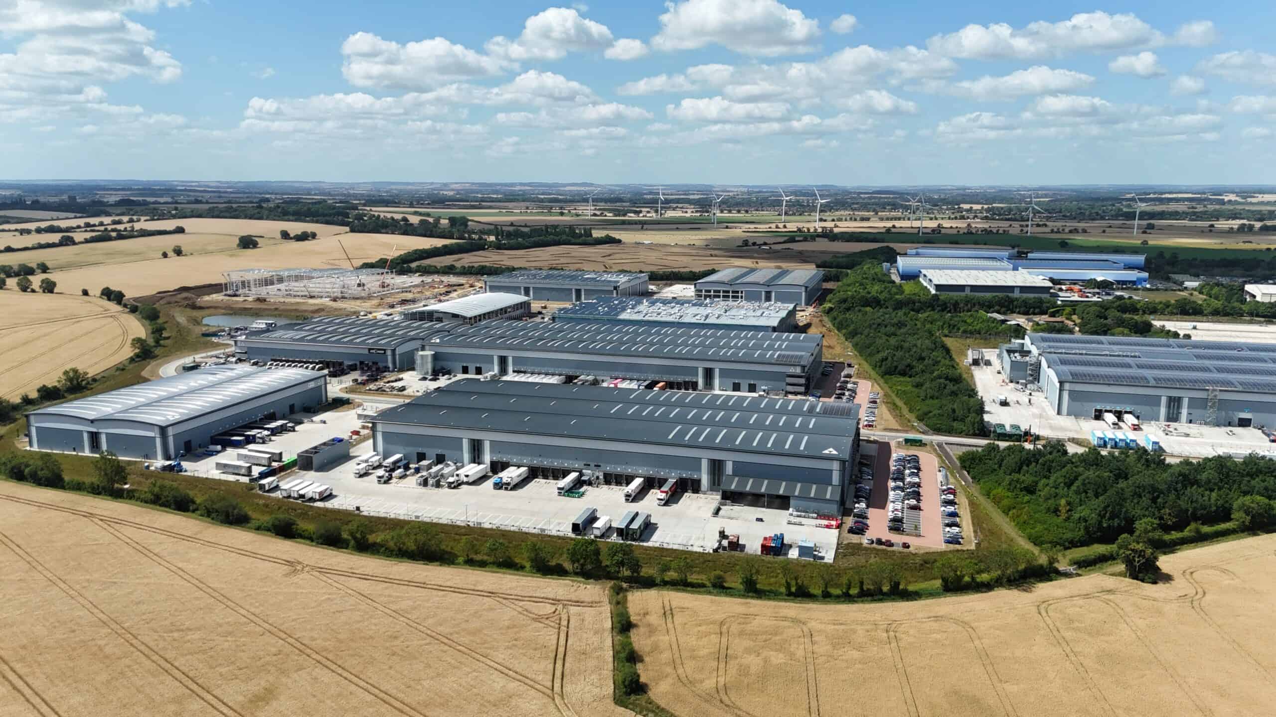 An aerial view of an industrial complex featuring several large warehouses with metal roofs. The surrounding landscape includes fields and small patches of green vegetation, with a distant view of power lines and a body of water in the background.