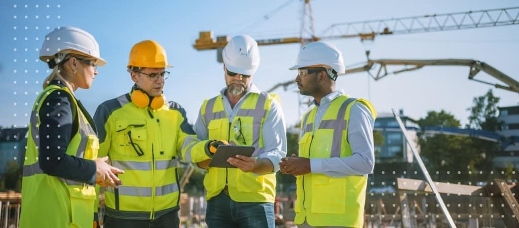 Construction team discussing project plans at a busy site with cranes and equipment.