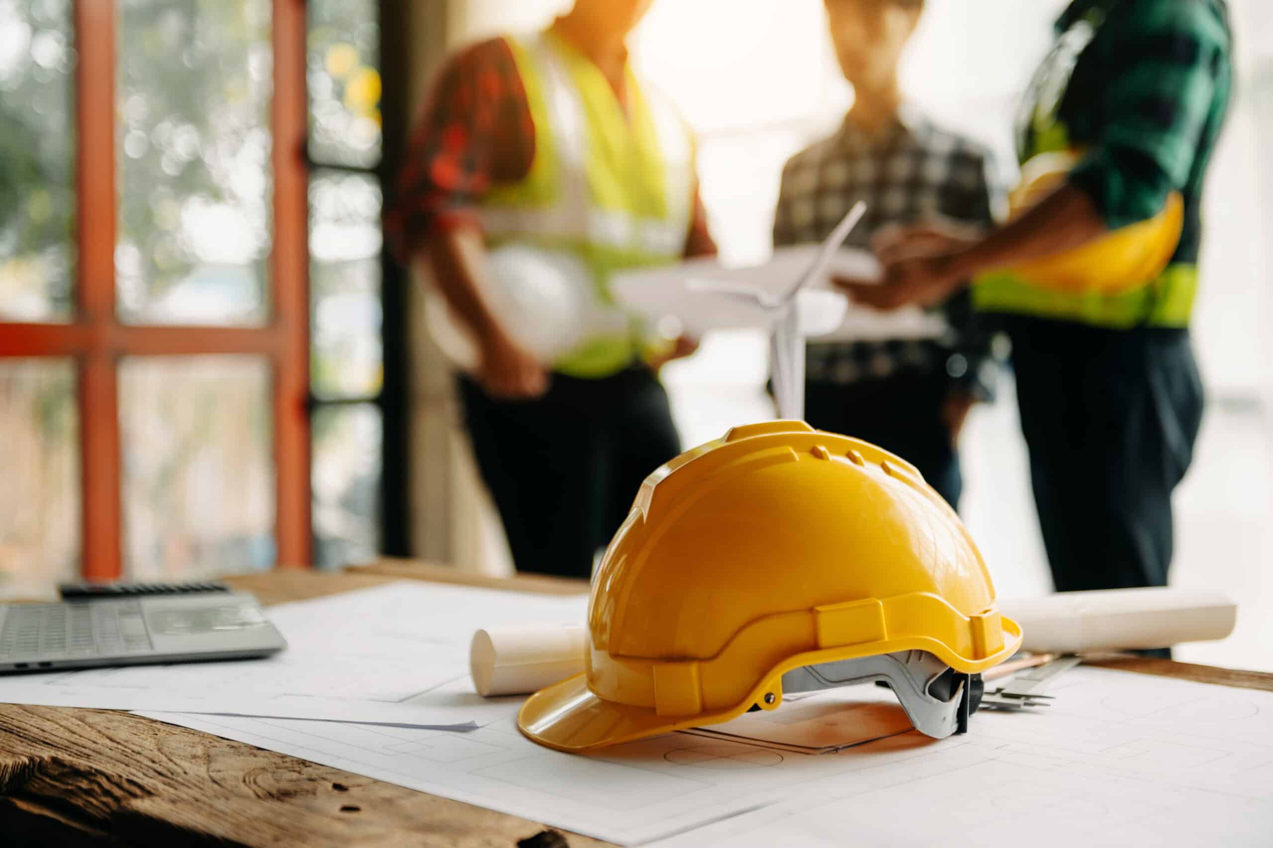 Construction safety helmet and blueprints on a table with contractors discussing plans.
