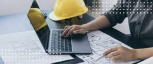 Hands type on a laptop resting on blueprints beside a hard hat, editing a construction contract in a document editor.
